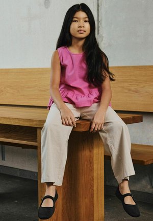 Pige med langt sort hår iført en lyserød ærmeløs top, beige bukser og sorte sko, der sidder på en træbænk mod en grå væg.