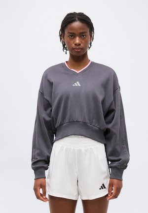 Moteris dėvi pilką ilgomis rankovėmis džemperį su rožine apdaila ir baltus sportinius šortus, stovinti priešais paprastą baltą foną.