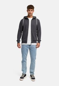 Mörkgrå zip-hoodie med dragsko i huvan, två framfickor, bärs över en vit t-shirt, ihop med ljusblå jeans och sneakers.