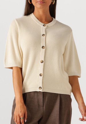 Korte mouw crèmekleurige gebreide cardigan met geribbelde textuur, knoopsluiting en een opstaande kraag. Gecombineerd met bruine op maat gemaakte broeken.