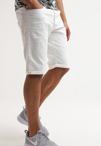 Shorts en denim blanc avec une coupe droite, un ourlet retroussé et des poches latérales. Portés avec des chaussures de sport grises et une chemise à motifs noir et blanc.