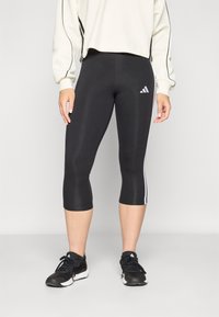 Juodos trumpintos leggingai, pagaminti iš tamprios medžiagos, su baltais šoninėmis juostelėmis ir „Adidas“ logotipu kairiajame šlaunyje.