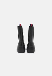Un par de botas de cuero negras hasta la mitad de la pantorrilla con suelas gruesas y lengüetas en rojo, blanco y azul, mostradas desde atrás sobre un fondo blanco.