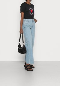Camiseta gráfica negra, jeans de pierna ancha azul claro, sandalias negras con tiras y un pequeño bolso negro con detalle de cadena.