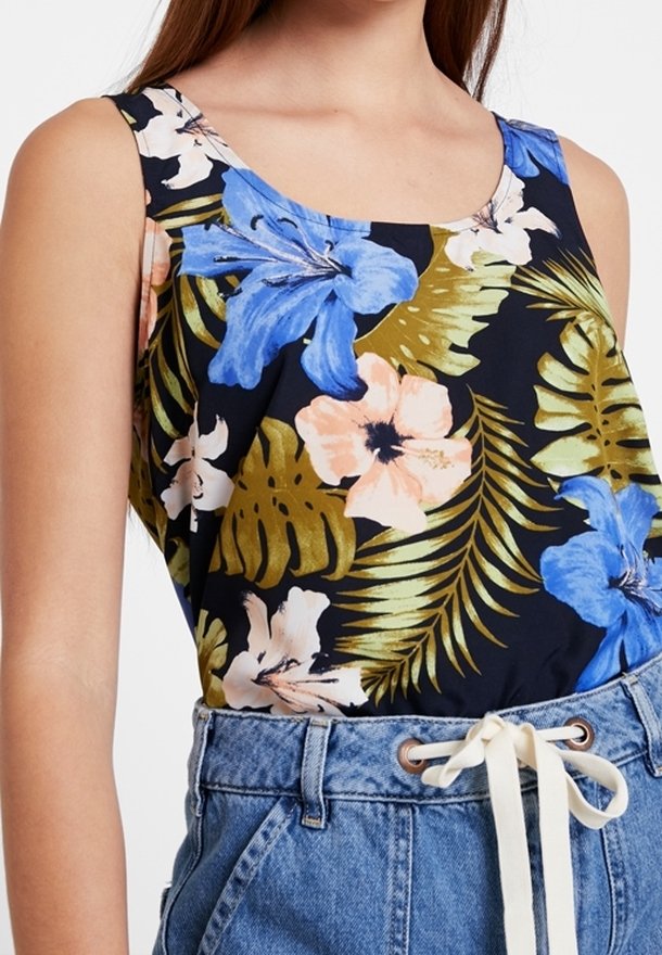 Blouse sans manches avec des fleurs tropicales bleues, roses et blanches et des feuilles de palmier vertes, associée à un jean taille haute avec une ceinture blanche à nouer.