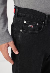 Black denim jeans with a textured finish, featuring metal hardware and a small logo patch on the front pocket. Gray sweater visible at the wrist.