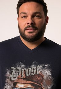 Námořnické tričko s výstřihem do V, které má grafický potisk s textem a portrétem, vyrobené z měkkého materiálu s hladkou texturou.