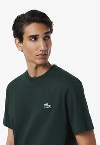 Maglietta in cotone verde scuro con scollo rotondo. Presenta un piccolo logo di coccodrillo ricamato in blu e bianco sul lato sinistro del petto.