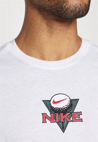 Balta medvilninė marškinėlė su apvalia iškirpte, kuriuose yra spalvingas išsiuvinėtas logotipas su golfu ir "NIKE" užrašas krūtinėje.