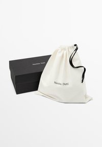 Katoenen draagkoordzak in crème met het "Massimo Dutti"-logo; daarnaast een zwarte doos met vergelijkbare branding; minimalistisch ontwerp, gladde textuur.