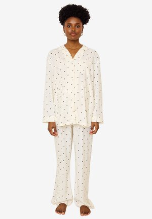 Woman standing barefoot wearing cream-colored long-sleeve button-up pajama set with small black heart patterns on top and pants.