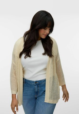 Woman with long dark hair wearing a cream open-knit cardigan over a white top and blue jeans, looking down to her left.