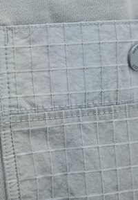 Lähikuva vaaleanharmaasta kangastaskusta, jossa on ruudukkoommel ja osittain näkyvä nappi teksturoidulla taustalla.