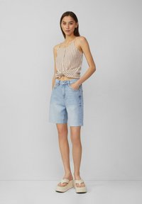 Haut cropped beige à rayures avec des boutons, assorti à un short en denim bleu clair avec un ourlet effiloché. Le mannequin se tient sur des sandales plates-formes.