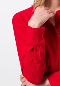 Chemise rouge à manches longues avec un poignet boutonné, en tissu lisse. Elle présente un col et une silhouette épurée, avec une coupe ajustée au poignet.