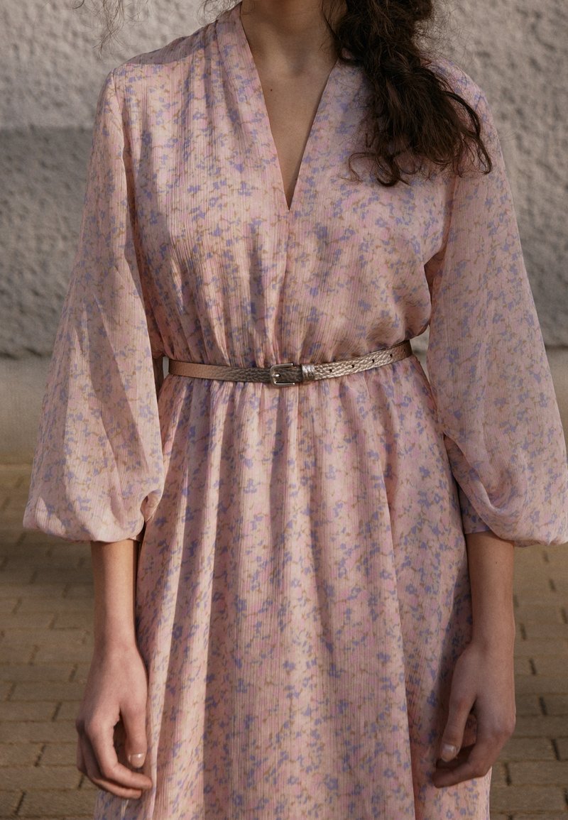 Femme portant une robe fleurie rose clair avec de longues manches bouffantes et une fine ceinture métallique à la taille, debout contre un mur texturé.
