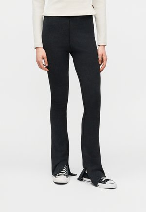 Sorte flarebuks med forreste slidser båret med sorte og hvide sneakers og en langærmet hvid top, model stående foran en ensfarvet baggrund.