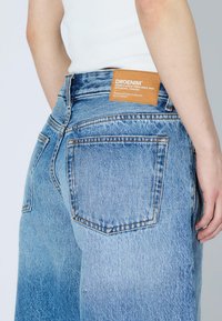 Jean en denim bleu clair avec une finition délavée, doté de poches arrière et d'une étiquette en cuir marron sur la ceinture, mettant en valeur un design décontracté.