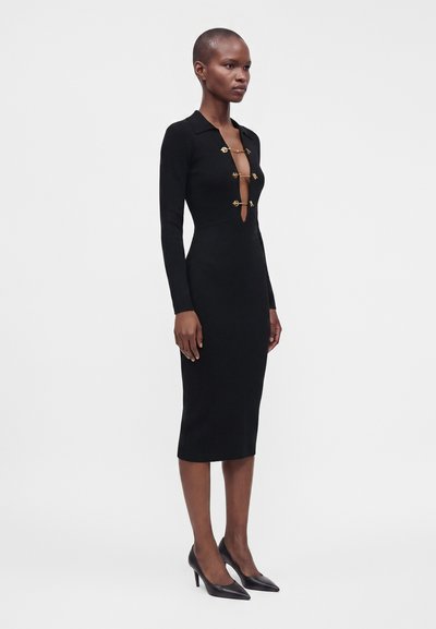 Femme portant une robe midi noire à manches longues avec trois fermetures en épingles de sûreté dorées sur un décolleté en V profond et des escarpins noirs à bout pointu.