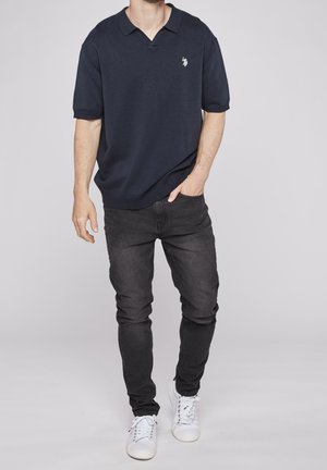 Polo azul marino de manga corta con cuello en V y logo blanco; combinado con jeans ajustados negros y zapatillas blancas, resaltando un look casual.