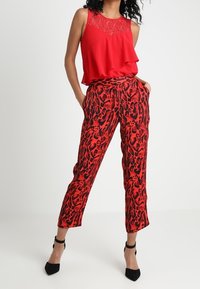 Haut sans manches rouge avec détail en dentelle, design en couches. Pantalon rouge et noir à motifs avec poches, coupe effilée et longueur cheville. Talons noirs.