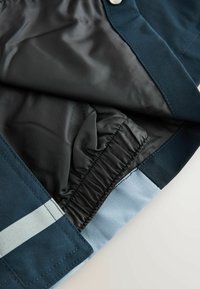 Nahaufnahme des Saums einer marineblauen Jacke mit elastischem Innenfutter und hellblauem reflektierenden Streifen auf dem äußeren Stoff.