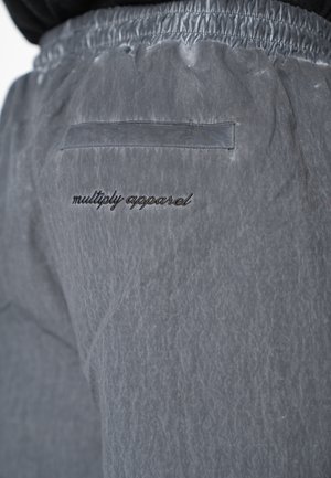 Prim-plan cu pantaloni din material gri, cu talie elastică și buzunar în spate, deasupra textului brodat negru „multiply apparel”.