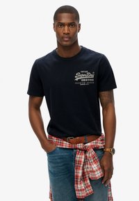 Marineblauw katoenen t-shirt met een ronde halslijn en een klein logo op de borst, gecombineerd met blauwe jeans en een rood geruit overhemd dat om de taille is gestrikt.