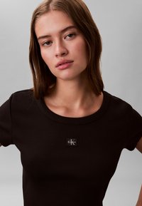 Noor daampruunide sirgete juustega noor naine kannab musta soonikutest t-särki, millel rinnal Calvin Kleini logo, halli tasase tausta ees.