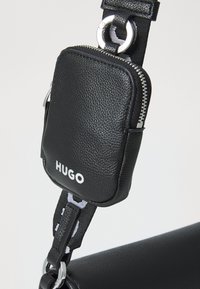 Svart lær crossbody-veske med teksturert overflate, utstyrt med en avtagbar glidelåspose og sølvfarget hardware. Logo vist på posen.