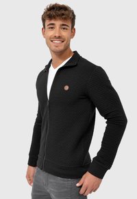 Sort zip-up jakke med tekstureret stof, ribbet manchetter og krave. Har et lille brunt logo patch på venstre bryst.