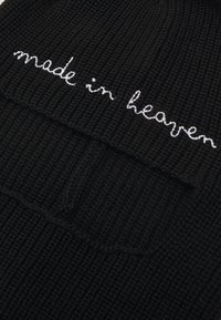 Zwarte gebreide beanie met een geribbelde textuur en witte geborduurde tekst "gemaakt in de hemel." Inclusief een voorzakdetail.