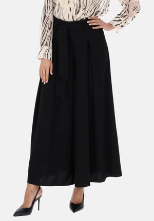 Donna che indossa pantaloni neri a vita alta e gamba larga con cintura annodata, abbinati a décolleté nere a punta e camicetta fantasia.