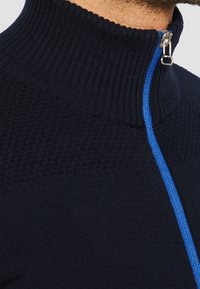 Marinblå zip-up tröja med hög ribbad krage. Har en kontrasterande blå dragkedja och ett texturerat stickmönster på den övre delen.