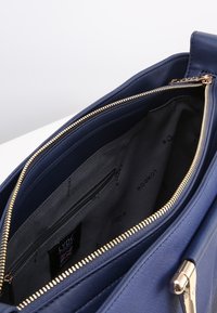 Námořnická modrá kabelka s texturovaným vnějším materiálem, zlatým zipem a prostorným černým interiérem s další kapsou na zip.