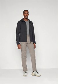Uomo in giacca grigia con zip, pantaloni cargo grigi chiari con zip e scarpe da trekking bianche su sfondo neutro.