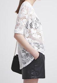 Weißes Spitzenoberteil mit floralen Ausschnitten und umgeschlagenen Ärmeln, kombiniert mit schwarzen Leder-Shorts. Kürzerer, lockerer Schnitt und zarte Textur. Schwarze Tasche mit Kettenriemen.