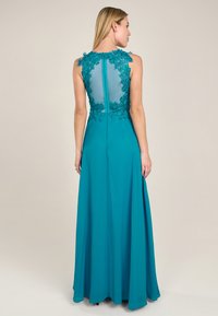 Türkise Abendkleid mit transparentem Rücken und floralen Spitzenakzenten. Der lange, fließende Rock verleiht Eleganz, mit einem zentralen Reißverschlussdetail.