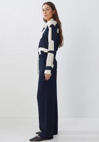 Navy wide-leg trousers paired with a patterned blouse featuring cream accents. Blouse has a collar and long sleeves. Model stands sideways.