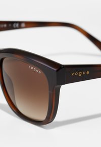 Ochelari de soare cu model țestoasă, cu o ramă pătrată, lentile maronii cu gradient și detalii aurii pe tempuri. Textură netedă din plastic.