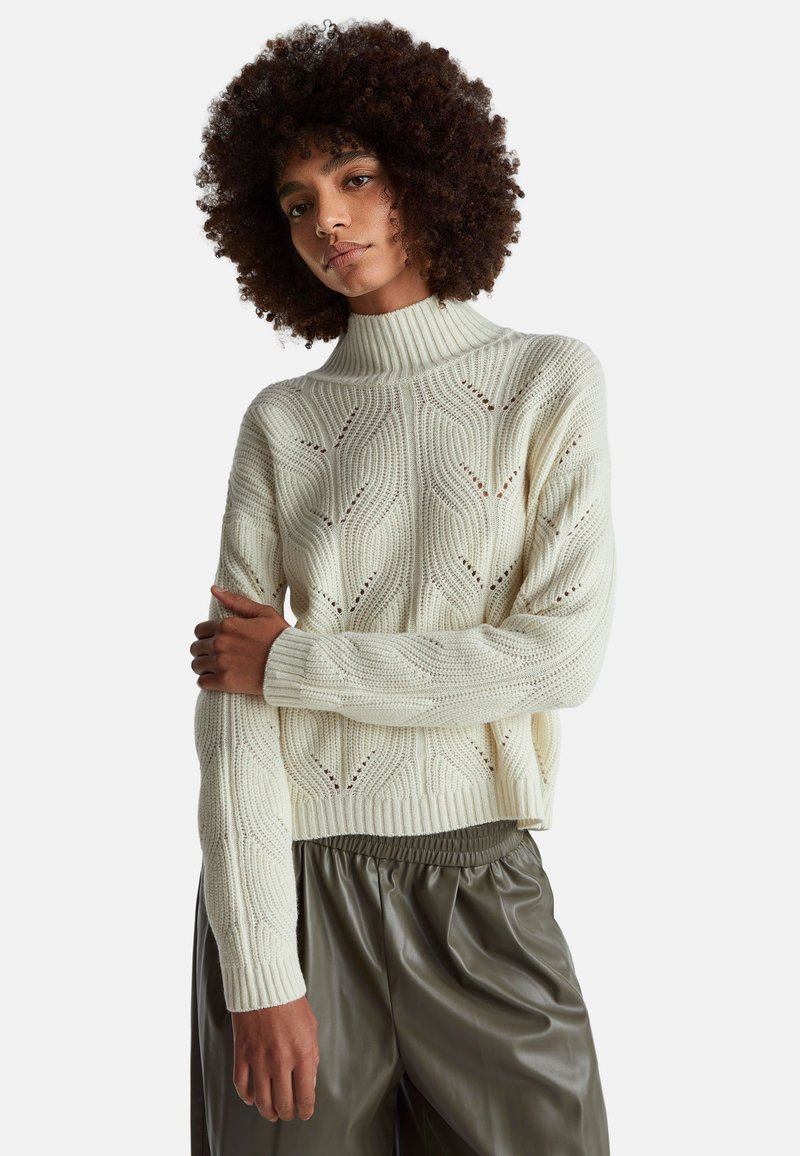 Suéter de punto color crema con un alto cuello acanalado, que presenta un patrón ondulado y detalles de puntada abierta, combinado con pantalones sueltos de color verde oscuro.