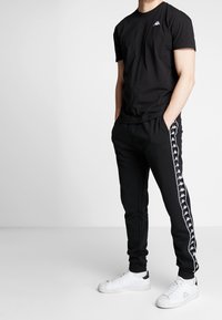 Černé tričko s krátkým rukávem a malým logem na hrudi spárované s černými zúženými tepláky s bílým postranním pruhem a bílými teniskami.