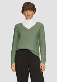 Groene V-hals trui met lange mouwen en ribbeltextuur, gecombineerd met een witte coltrui eronder en zwarte broek.