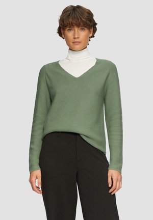 Grüner V-Ausschnitt-Pullover mit langen Ärmeln und gerippter Textur, kombiniert mit einem weißen Rollkragenpullover darunter und schwarzen Hosen.