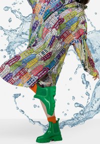 Kleurrijke, patroonrijke regenjas met huizen in verschillende tinten; gecombineerd met felgroene, glanzende enkellaarsjes en oranje sokken. Water spat rond.