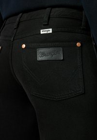 Gros plan de la poche arrière d'un jean Wrangler noir avec logo en patch en cuir et détails à rivets sur tissu foncé.
