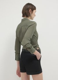 Camicia verde oliva in tessuto texturizzato con polsini a bottone, abbinata a pantaloni corti neri con cintura intrecciata e orlo arrotolato.