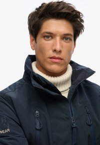 Marine waterdichte jas met hoge kraag, rits en trekkoorddetails. Binnenste ribgebreide trui in lichtroom. Glad, elegant stofafwerking.