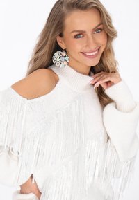 Witte gebreide trui met koud-schouderdesign, franjes en een gestructureerd patroon. Ronde, kleurrijke oorbellen zorgen voor contrast.