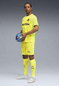 Uniforme da calcio giallo brillante con un motivo ondulato, abbinato a calze coordinate e scarpe da calcio grigie. Il giocatore tiene in mano un pallone da calcio blu.